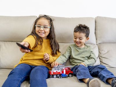 Sister and brother watching TV and playing with a firetruck on the couch.