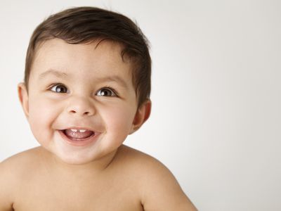 Baby boy with brown hair brown eyes smiling with two teeth showing, looking past the camera with no shirt on