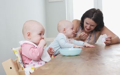 一对双胞胎女婴坐在餐桌旁，一个在用勺子喂，另一个在嚼她的拳头