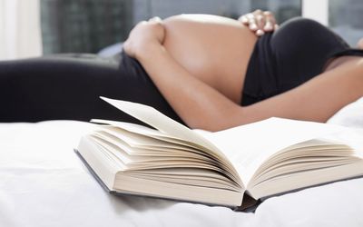 Mid section of pregnant woman lying in bed with open book in foreground