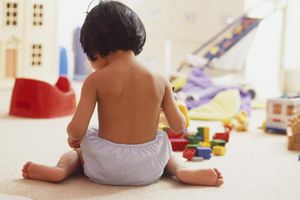 后视图的黑头发女孩蹒跚学步穿着训练裤坐在地板上，周围的玩具