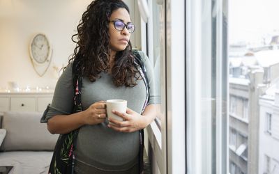 pregnant woman looking out window drinking coffee