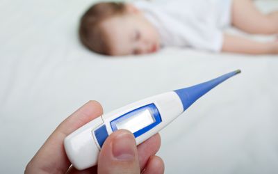 Parent holding a baby thermometer with a baby on a bed