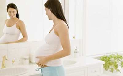 Pregnant woman with hands on stomach looking in mirror.