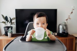 一个亚洲婴儿坐在高脚椅上，双手端着一个透明的吸管杯。