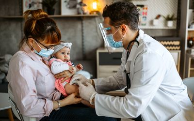 A mom wearing a mask holds her baby as a doctor gives her a vaccine in her leg.