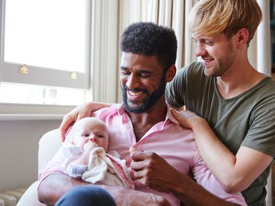 Loving Male Same Sex Couple Cuddling Baby Daughter On Sofa At Home Together