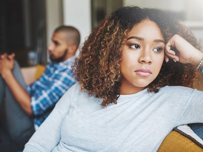 Woman sitting on couch with hand on head, man in background behind her