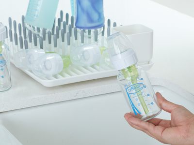 Hand holding Dr. Brown's Natural Flow Options+ Narrow Glass Baby Bottle over a sink next to a dish drying rack with other baby bottles