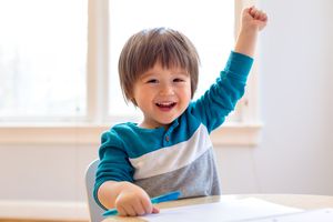 蹒跚学步的孩子微笑和举起他的手在桌子上，同时用他的另一只手握笔