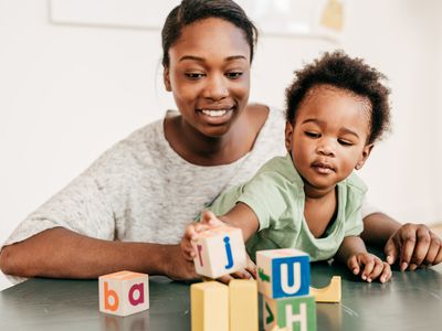 Person holding a child on their lap while they stack wooden alphabet blocks on a hard surface