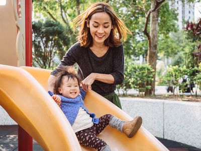 Young mother holding her toddler's hand as she goes down a yellow slide.