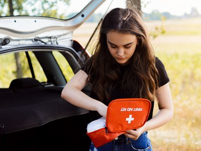Young attractive girl stands near car with open back door and emergency lights is on, tries to find something in first aid kit