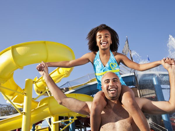 A young girl in a blue bikini smiles and sits on her father's shoulders at a water park.