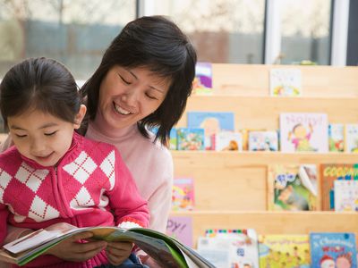 Girl (4-5) sitting on lap of teacher reading book, smiling