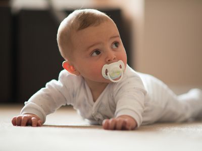 Baby laying on the floor wearing a white onesie and pacifier against a blurred background