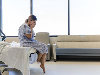 A woman in a hospital gown sits on the edge of a hospital bed, head in hands.
